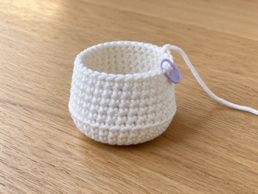 White crochet nurse uniform base in progress with a rounded bottom, defined lower ridge, and purple stitch marker.